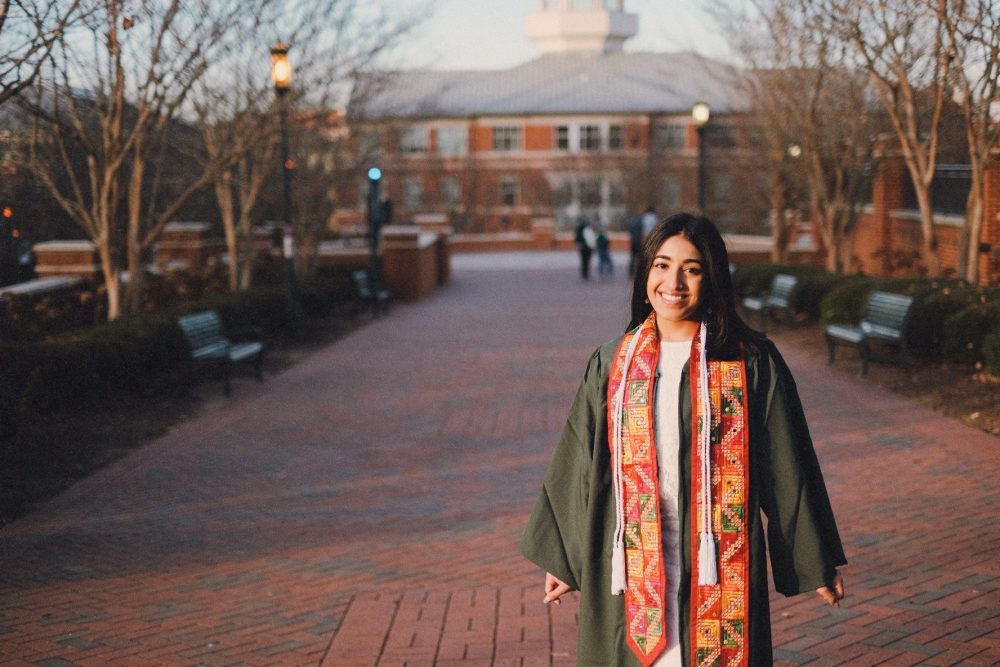 Young woman standing on UNC Charlotte campus in green graduation robe and regalia.