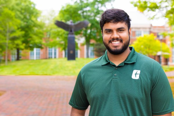 John Gopali standing on campus at UNC Charlotte with eagle statue in Woodward Hall quad in the background